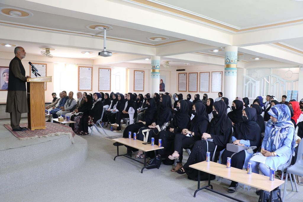 women sitting in chairs, dressed all in black, listening to a lecture given by a man standing on a platform behind a podium while the world watches