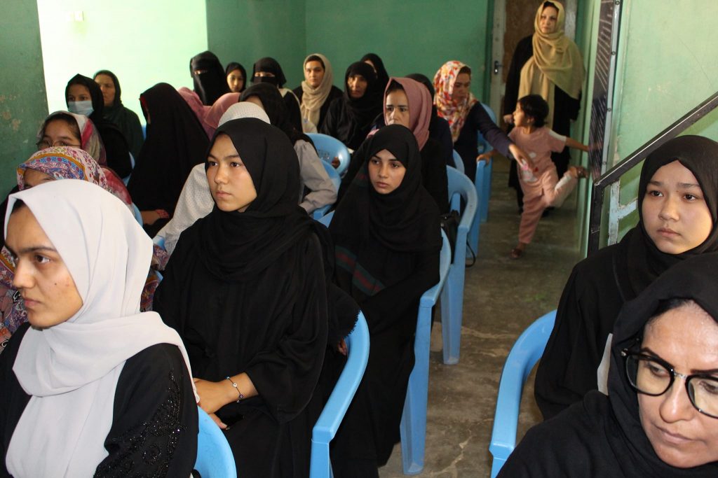 Women sitting in chairs, mostly dressed in black robes and headscarves, listening to a speaker